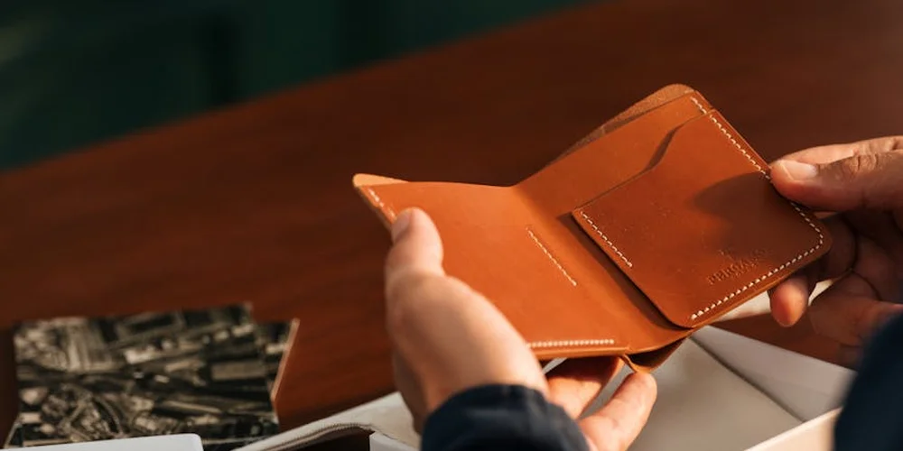 A person holding an open brown leather wallet over a wooden table.
