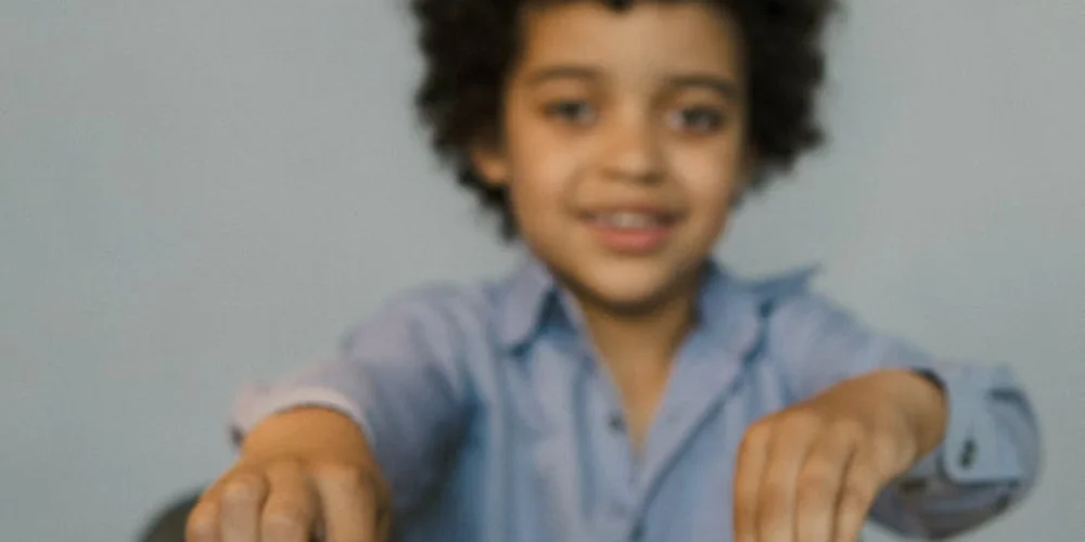 A young child with curly hair wearing a light blue shirt smiles at the camera indoors.