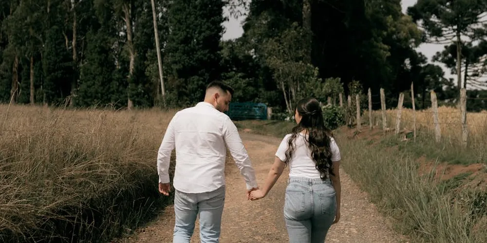 A man and a woman hold hands while walking on a dirt path through a grassy field, with trees in the background.