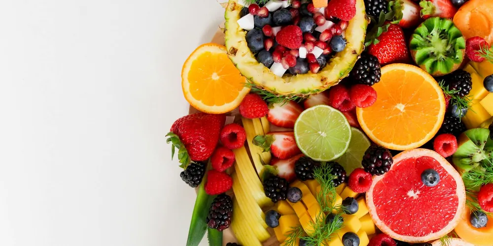Assorted fresh fruits arranged in a colorful display on a white background, including orange slices, lime halves, pineapple chunks, strawberries, raspberries, blueberries, blackberries, and other fruit pieces.