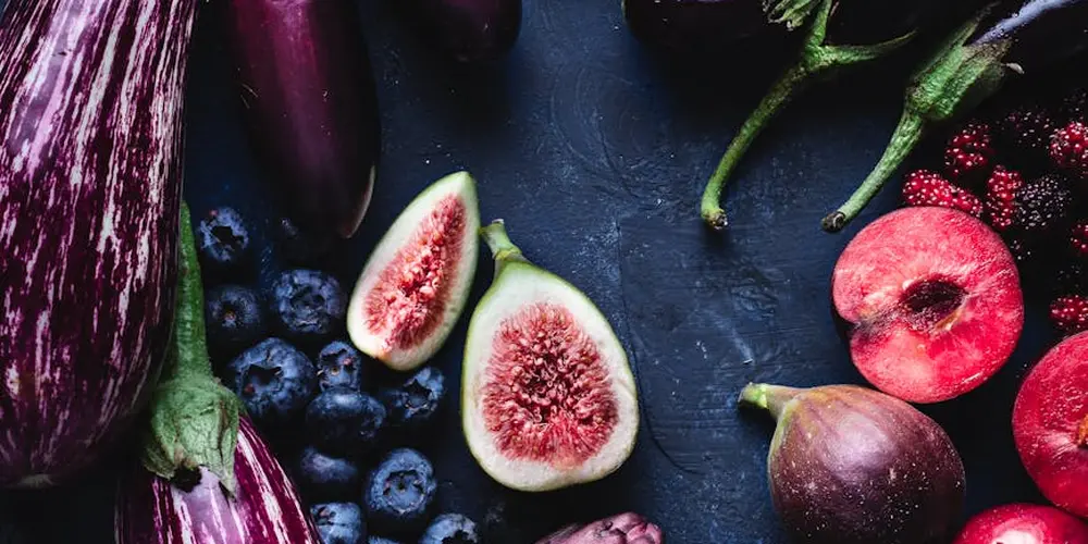 Assorted fruits and vegetables arranged on a dark surface, including figs, blueberries, plums, and eggplant