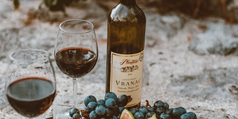 A wine bottle, two glasses of red wine, and a small cluster of grapes on a rough stone surface.