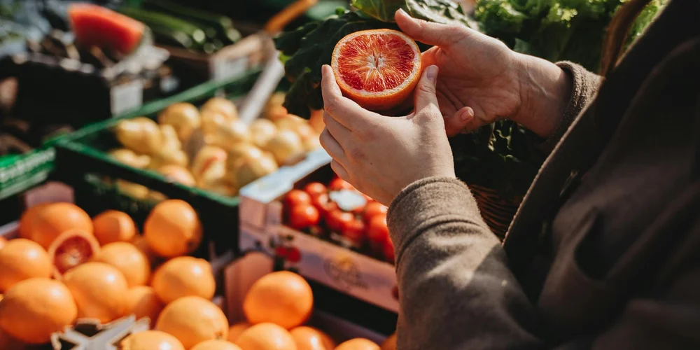 Hands hold a halved citrus fruit at a market with baskets of fresh produce in the background.