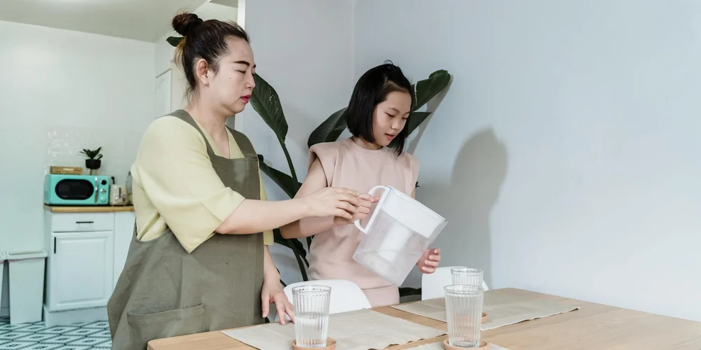 Two women in a kitchen handle a white pitcher and a glass, preparing water at a table.
