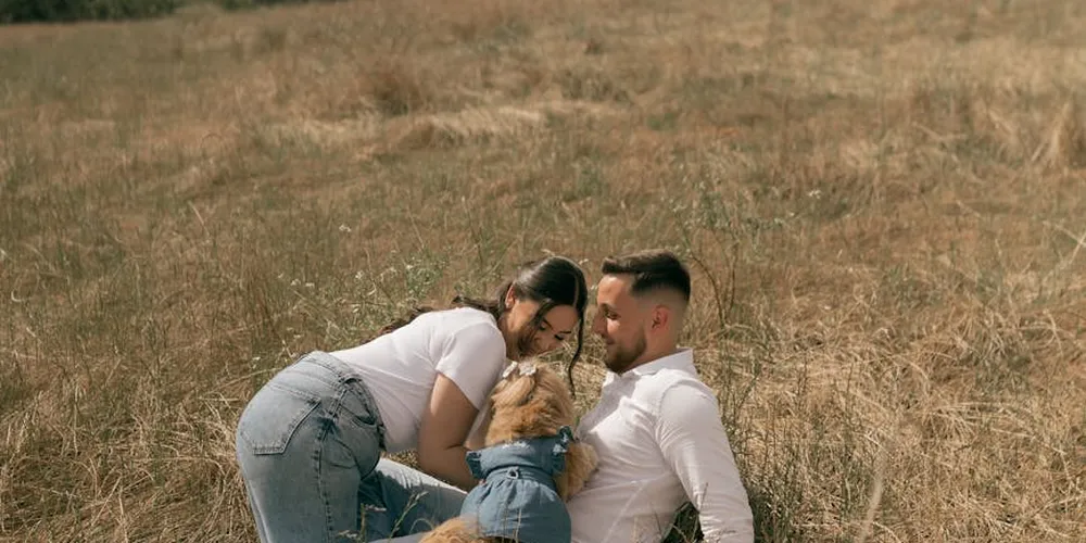 Two adults crouch in a sunlit grassy field, interacting with a small fluffy dog.