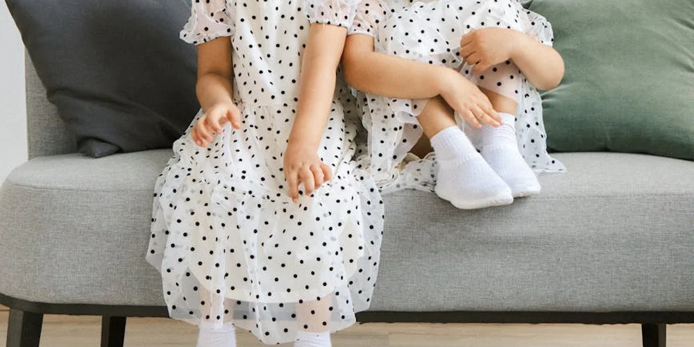 Two children wearing white polka-dot dresses sit on a light gray sofa.