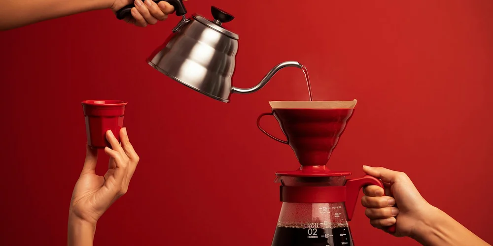 Two hands pouring water from a kettle into a red pour-over coffee maker against a red background.