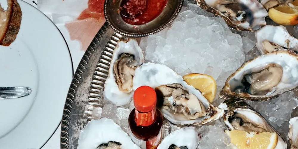 Oysters on ice with lemon wedges and a small red bottle of sauce on a silver tray, with a white plate and cutlery in the background.