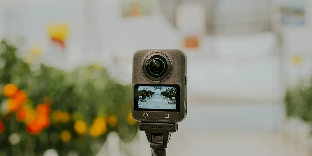 Close-up of a small action camera mounted on a stand with a blurred greenhouse or indoor garden in the background.