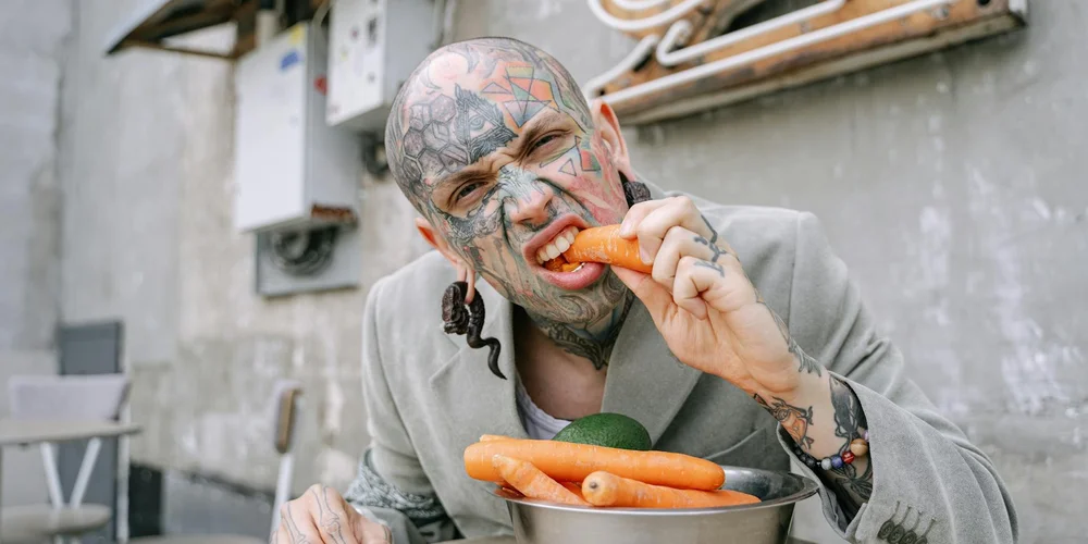 A tattooed man in a gray hoodie bites into a carrot while sitting outdoors, showcasing carrot as a healthy snack.