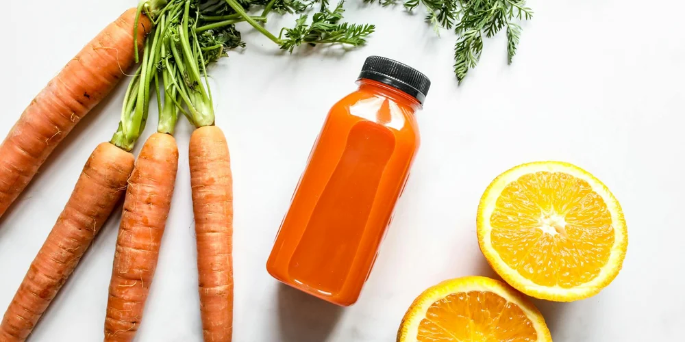 Fresh carrots with green tops, a bottle of orange juice, and sliced oranges on a white surface.