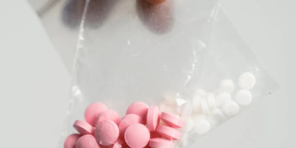 Close-up of pink circular tablets and white capsules in small clear plastic bags on a light gray surface.
