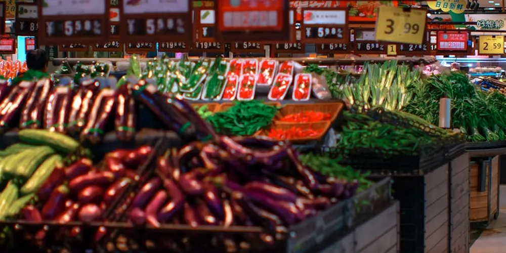 Market display of fresh vegetables, including eggplants and peppers, with leafy greens in the background.