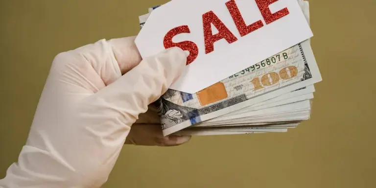 A gloved hand holding a fan of cash with a large red SALE tag against a plain background