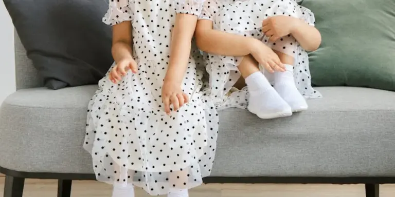 Two young children wearing white polka-dot dresses sit on a gray sofa.