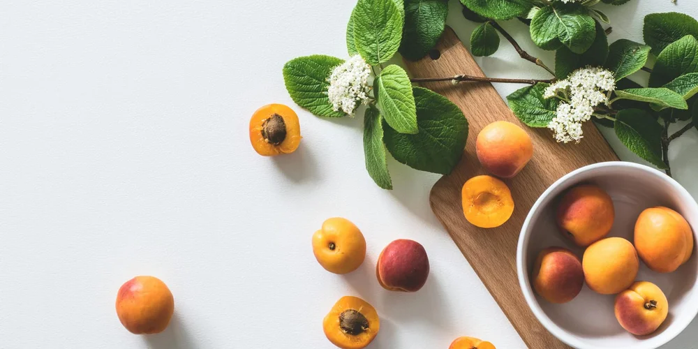 Fresh apricots and green leaves arranged on a white surface with a wooden cutting board and a bowl of apricots nearby.