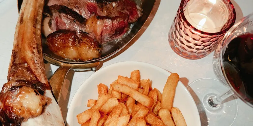 Roasted meat and French fries on a white plate with a skillet of meat nearby, a glass of red wine, and a lit candle in the background.