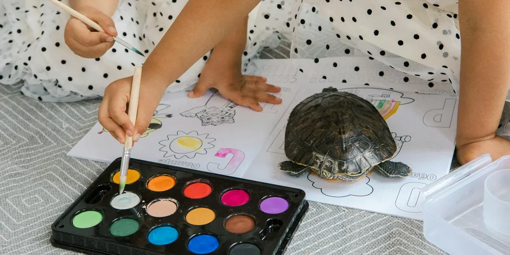Two children color a turtle drawing while a real turtle sits on the paper nearby.