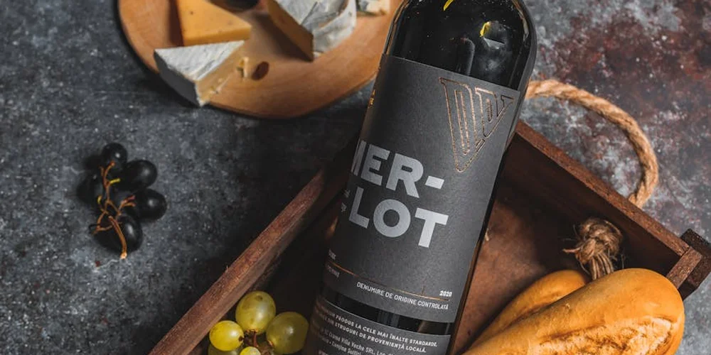 Bottle of Merlot wine on a dark countertop next to a small wooden board with green and black grapes.