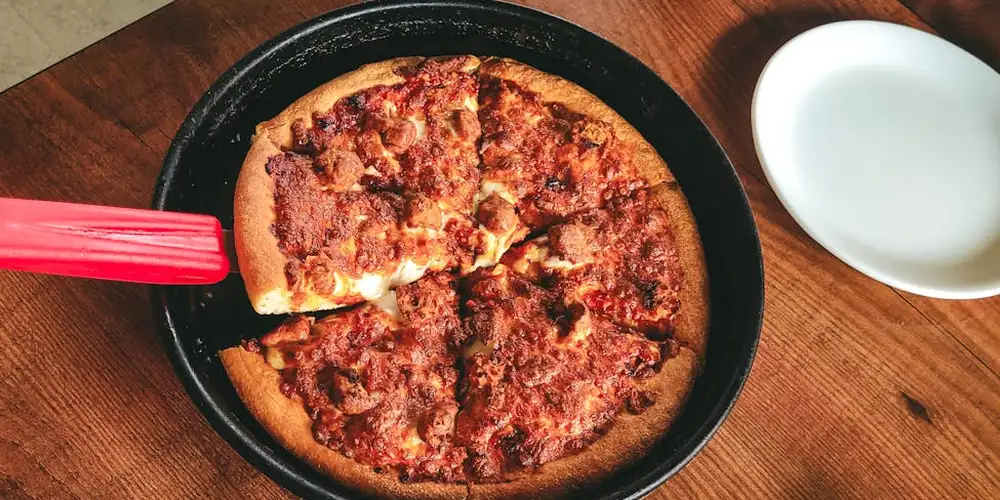 Pepperoni pizza in a black skillet on a wooden table, with a red spatula resting on the edge and a white bowl nearby.
