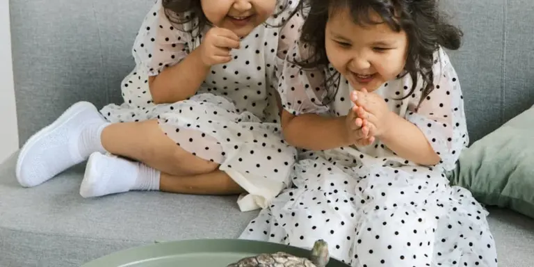 Two young girls wearing white polka-dot dresses sit on a gray couch, smiling and looking at something in front of them.