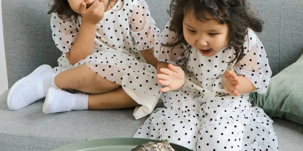 Two young children wearing white dresses with black polka dots sit on a gray couch, looking down at their hands.