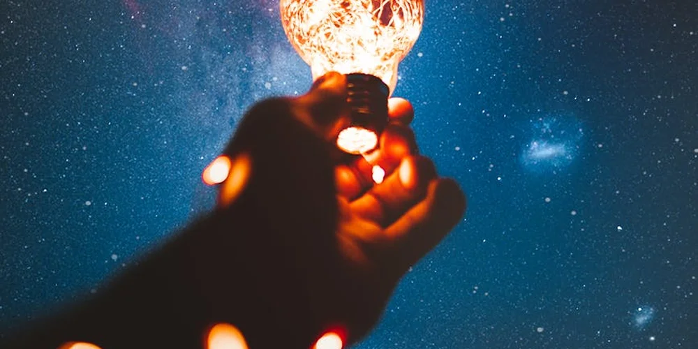 Hand holding a glowing light bulb against a starry night sky.