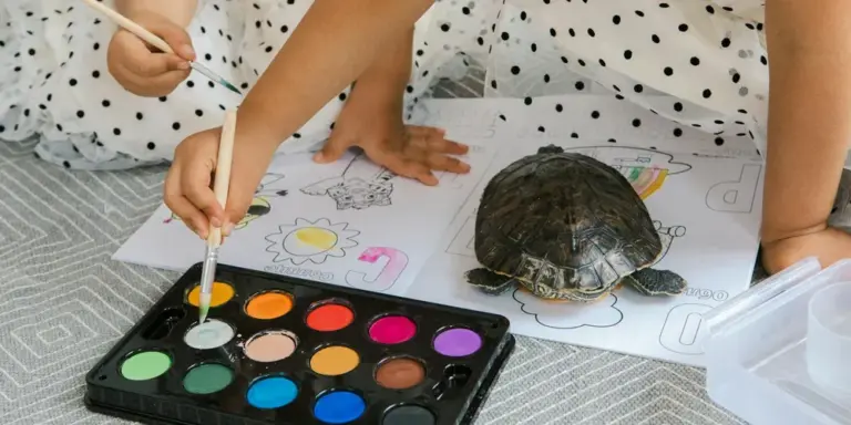 A pet water turtle sits on a drawing sheet while children color with pencils; a watercolor palette is in the foreground.