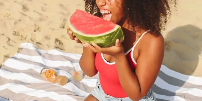 Woman on a sandy beach wearing a red swimsuit, biting into a large slice of watermelon held with both hands over a striped towel.