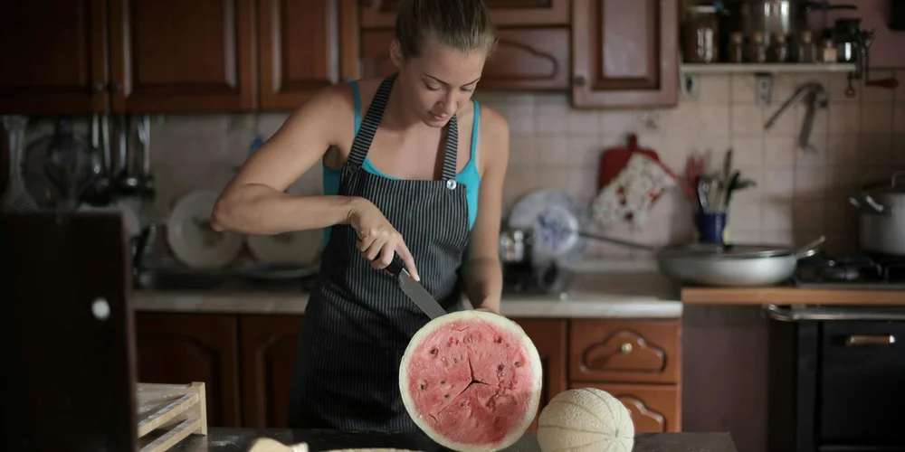 A person in a kitchen wearing an apron slices a watermelon with a knife, suggesting how to prepare watermelon for turtle feeding.