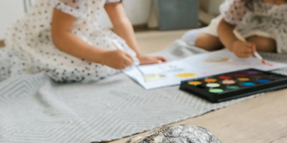 Two young children sit on a light-colored rug, drawing with markers and a paint set nearby.