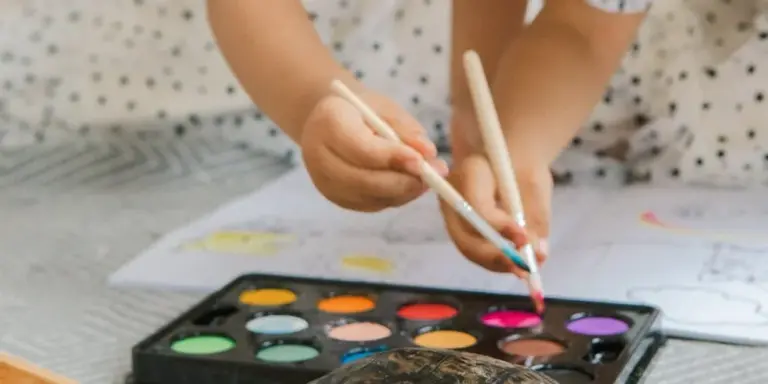 A child's hands hold paintbrushes over a watercolor palette with vibrant paints on a table.